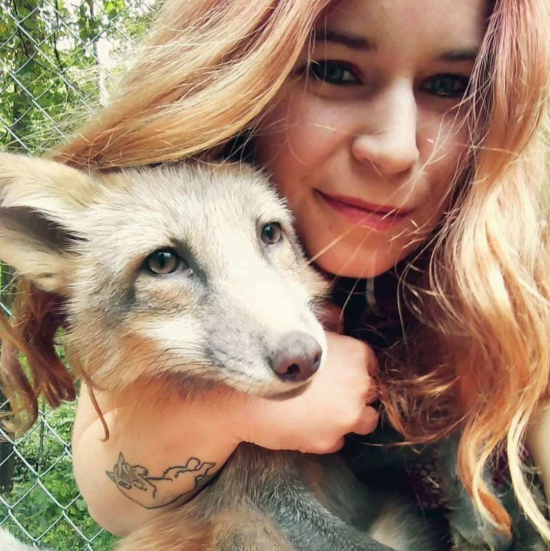 A photo of a woman with blonde hair smiling into the camera and holding a pet red fox